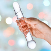 A hand holds the hypoallergenic Gläs 7" Callisto Clear Glass Dildo, a transparent cylindrical object with a rounded tip and base, set against a blurred background with soft, colorful lights.